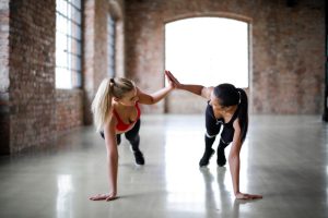 Fit smiling women in sportswear looking at each other and giving high five while doing push up exercise on gray glossy floor against blurred interior of spacious workout room with brick walls and big windows